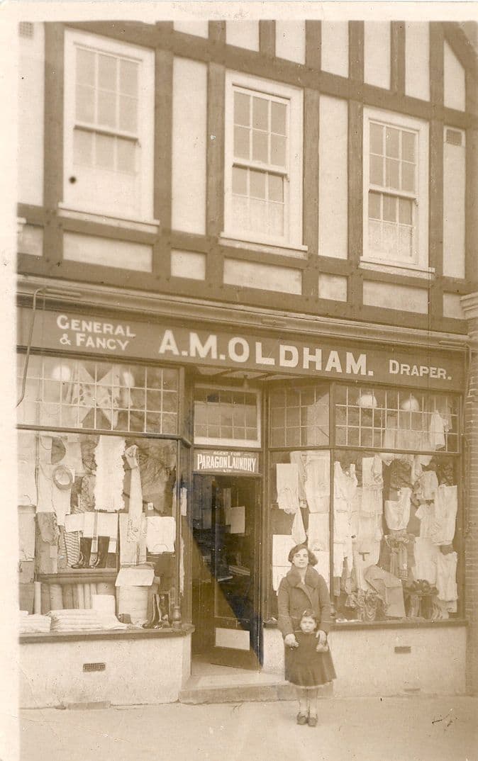 A mock-Tudor shop front with the fascia “General & Fancy A. M. Oldham Draper” and windows full of clothes and linens; a woman stands in the doorway with a small girl in front of her. A sign over the door reads “Agent for Paragon Laundry”.