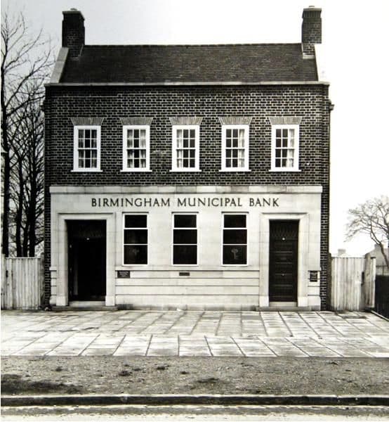 A newly built two-storey brick bank with sash windows, chimneys at each end and a pale stone ground floor lettered “Birmingham Municipal Bank”, with two doors, a paved forecourt and a wooden fence on either side.