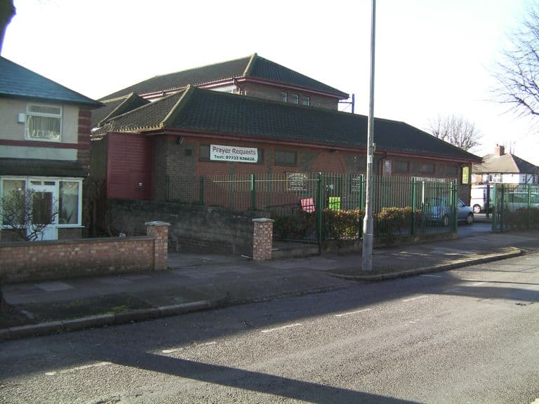 The low brick Methodist church buildings with tiled hipped roofs behind green railings, seen across the street beside a pebble-dashed house.