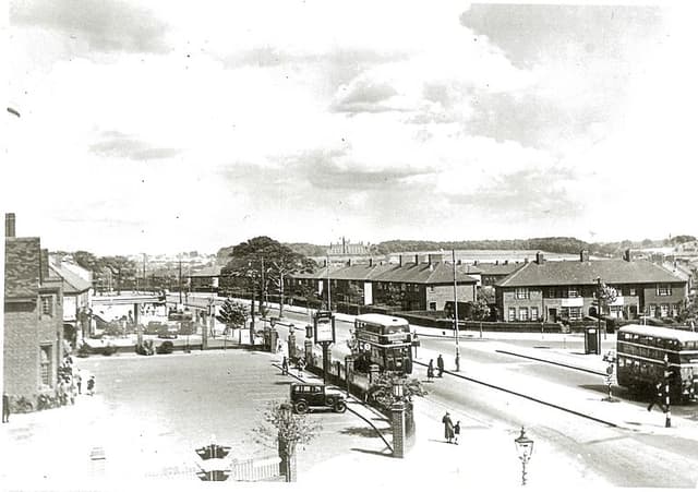 A raised view along College Road: the Crossways pub’s gate piers, hanging sign and forecourt on the left with a parked car, two double-decker buses and pedestrians on the road, rows of new brick houses across the junction, and open country and a large building on the skyline.