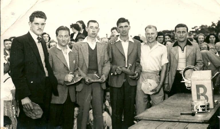 Six men in jackets and open-necked shirts stand in a row holding small cake stands, with a crowd behind them and a prize table with a lettered “B” board and trophies at the right.