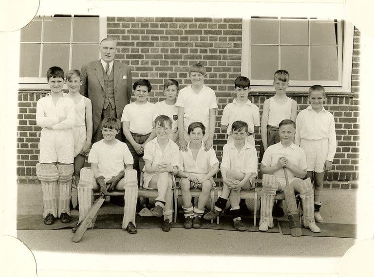 School cricket team of about thirteen boys in whites, two wearing pads and holding bats, with an older man in a three-piece suit standing behind.