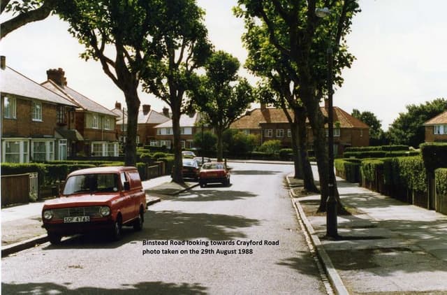 Colour photo of a tree-lined Binstead Road curving between brick semi-detached houses with hedges, a red Mini van parked on the left; a caption is printed on the image.