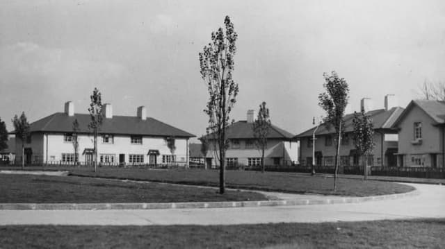 Newly built pairs of white rendered houses with tall chimneys set around a curving green planted with young trees.