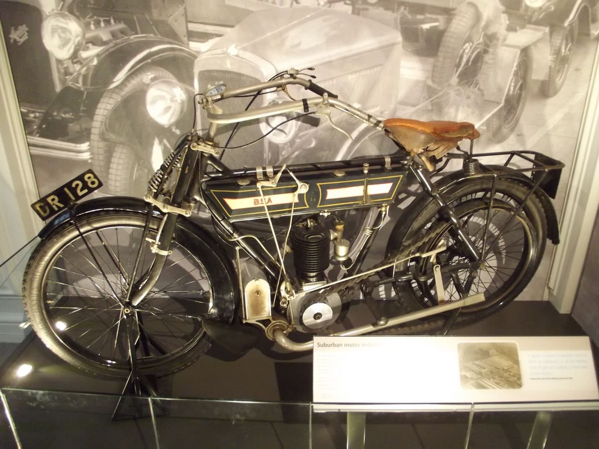 An early black motorcycle with a leather saddle and a single-cylinder engine in a museum display case.