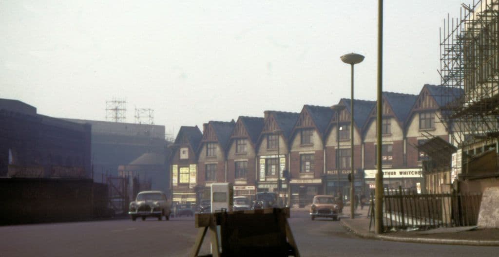 1960s street of old gabled shops beside the ruined walls of a large building, with a tower under construction.