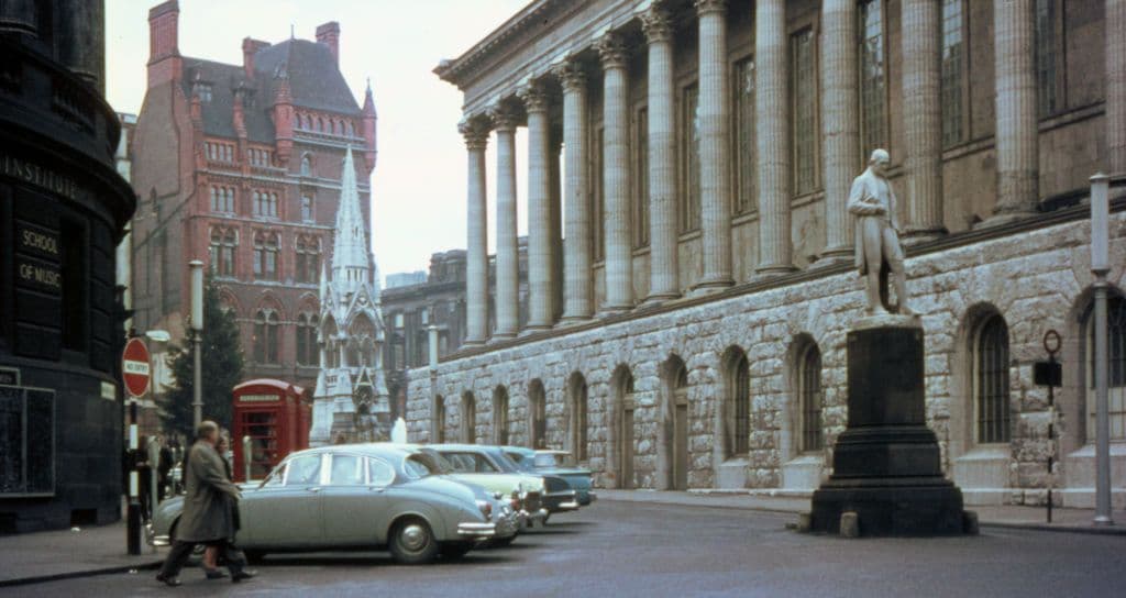 A 1960s street scene with a statue on a plinth in the foreground and a Gothic spire behind.