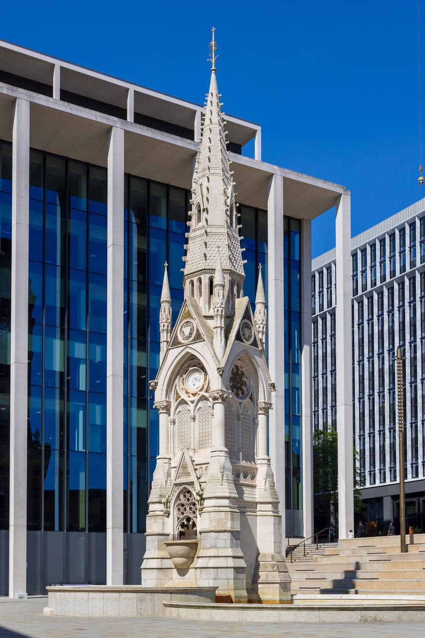 A Gothic stone spire with mosaic panels rising above a fountain in a city square.