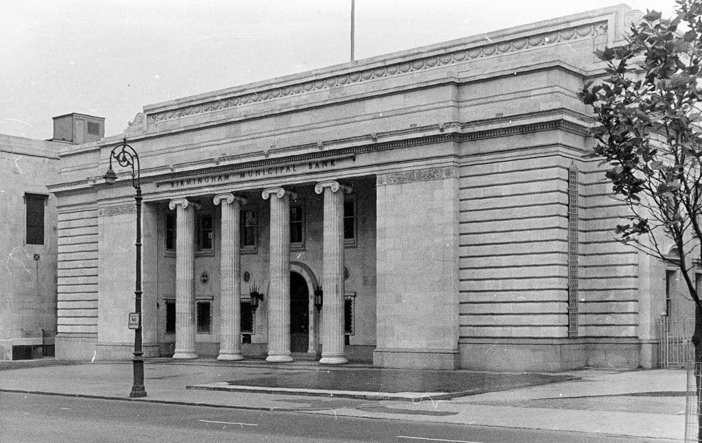 Black and white photograph of a pale stone classical building with tall columns across its recessed front, on a quiet city street.