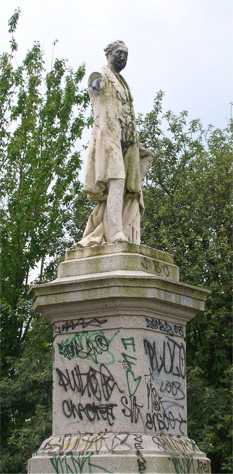 A white marble statue of a man with one arm outstretched, on a stone pedestal covered in graffiti, among trees.