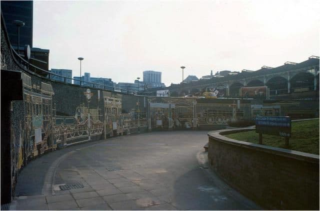 A sunken landscaped roundabout with a pedestrian walkway and mural, and a large station roof beyond.