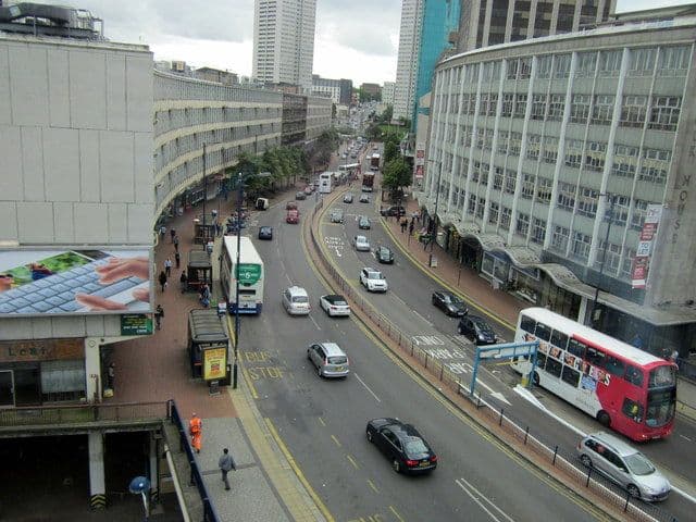 Elevated view along a busy dual carriageway with buses, lined by a long curving 1960s office block on the left and a curved building on the right.