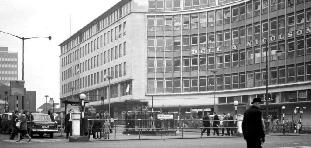 Black-and-white street photograph of a road junction with railings round the entrance to underground toilets and a large curved modern office block behind.