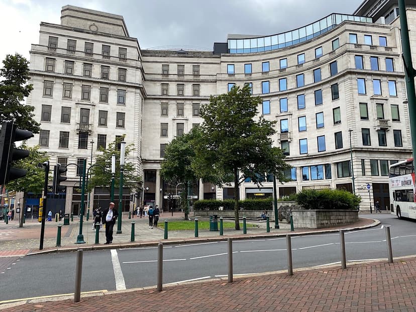 A small paved square with trees and bollards in front of a large curved Portland stone building.