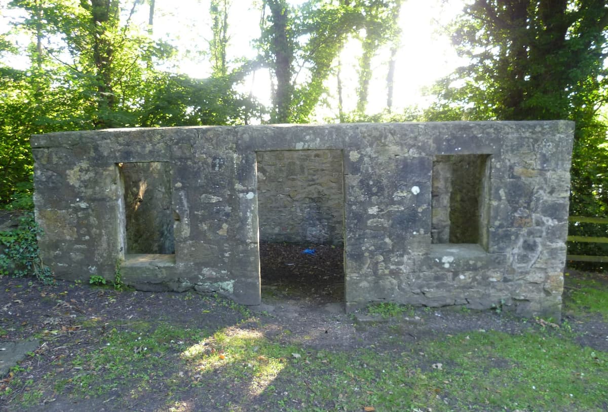 Roofless ruin of a small stone outbuilding among trees, with an old iron engine cylinder beside it.