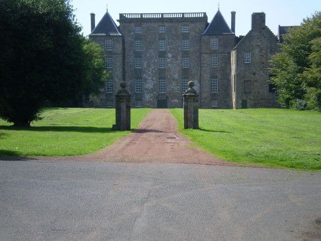A tall grey stone mansion with steep-roofed corner turrets and a parapet, seen up a drive between two stone gate piers.
