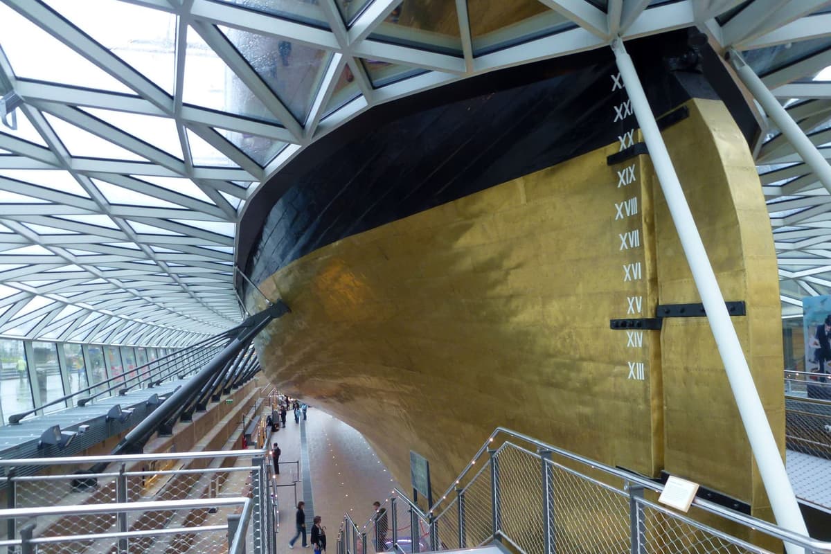 The coppery-gold metal-sheathed stern and rudder of a sailing ship, raised above a dry dock under a glass roof.