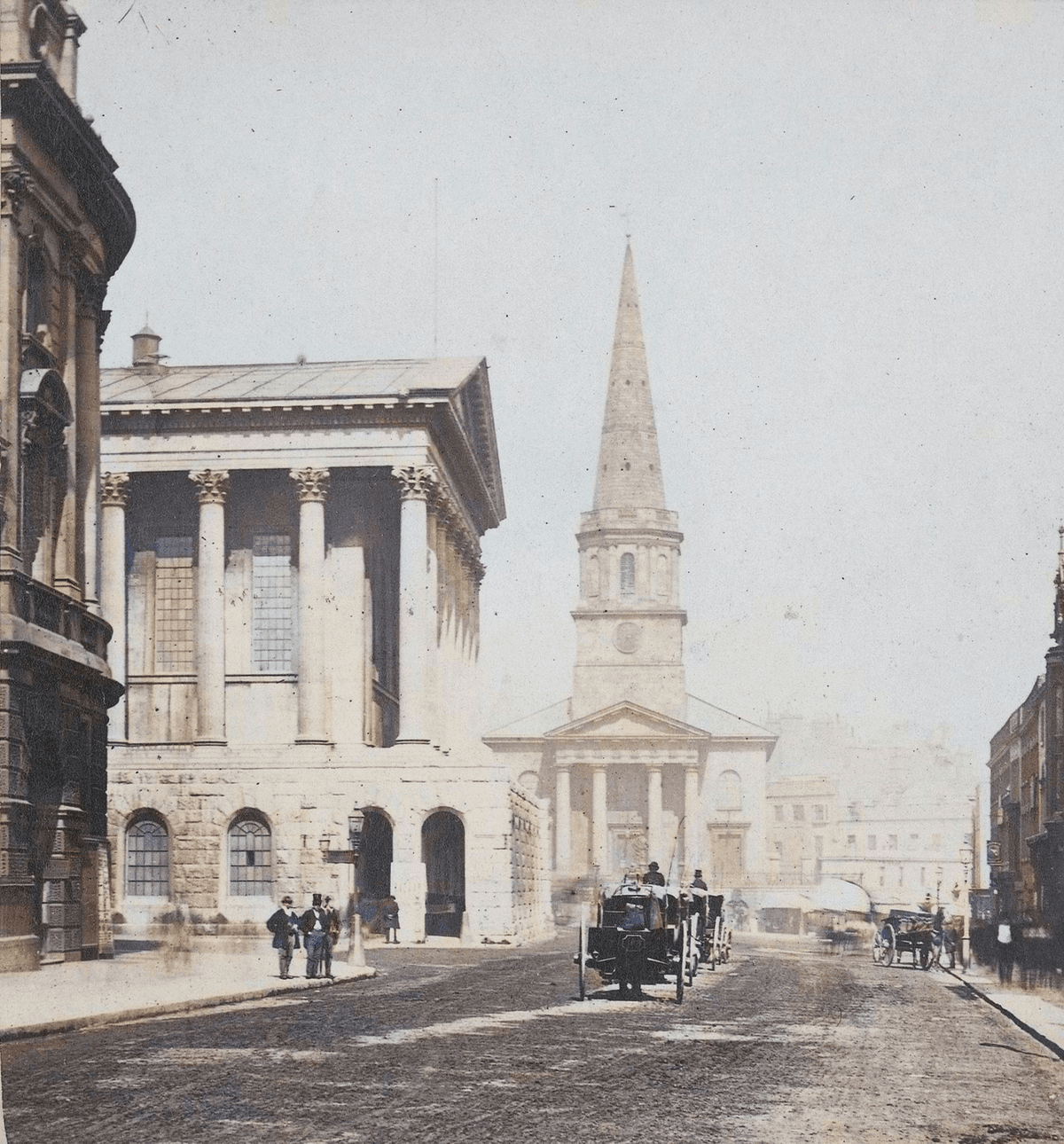 Victorian street photograph, artificially coloured, looking up a street past a columned hall to a church portico and spire.