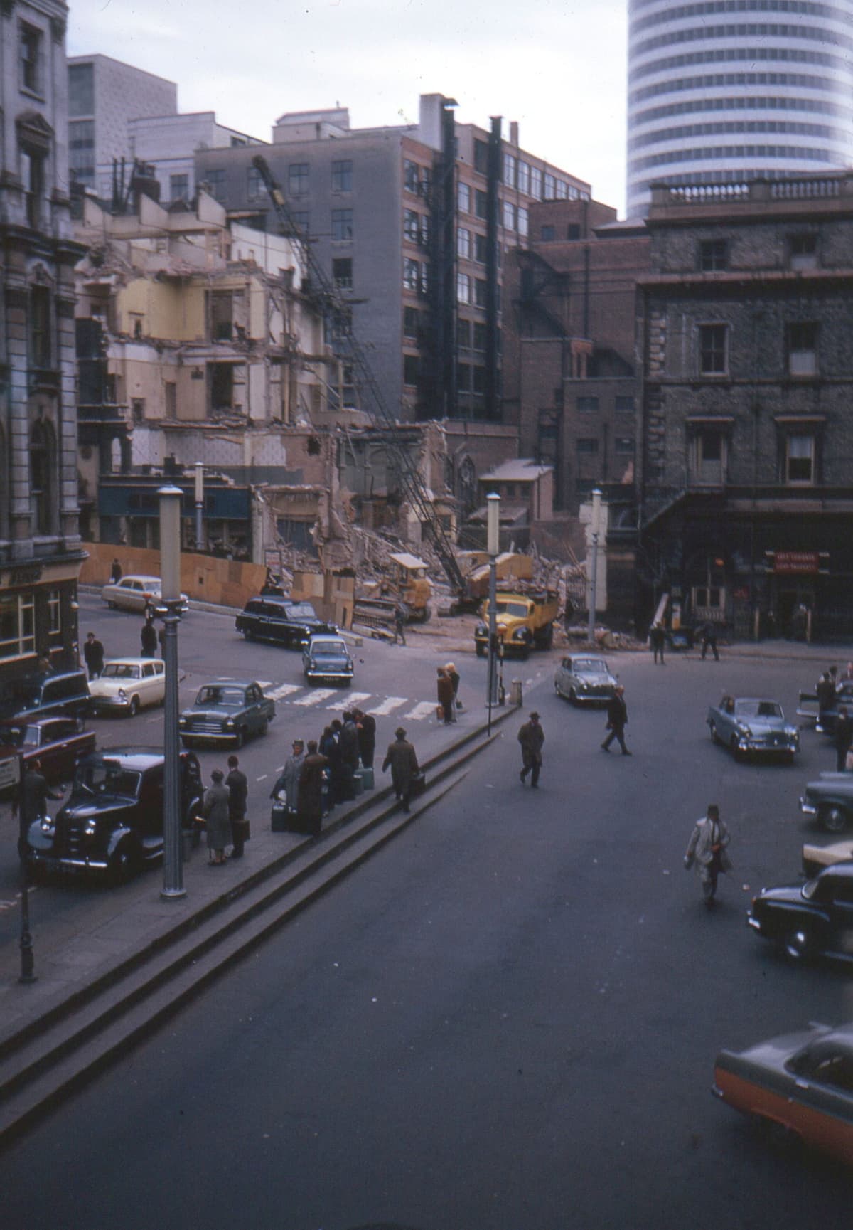Colour photograph of a Victorian building half demolished, with taxis waiting below and a new round tower block behind.