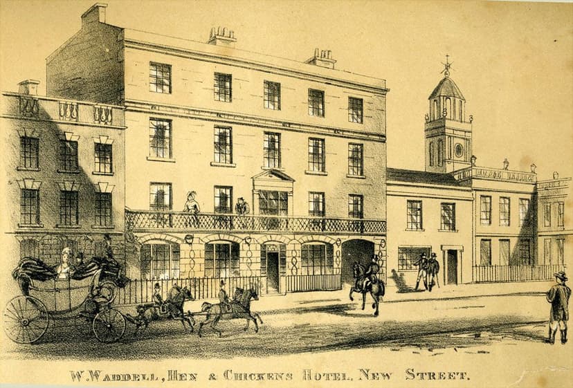 Print of a hotel front with a pillared portico over the pavement, figures on the balcony and a carriage arriving.