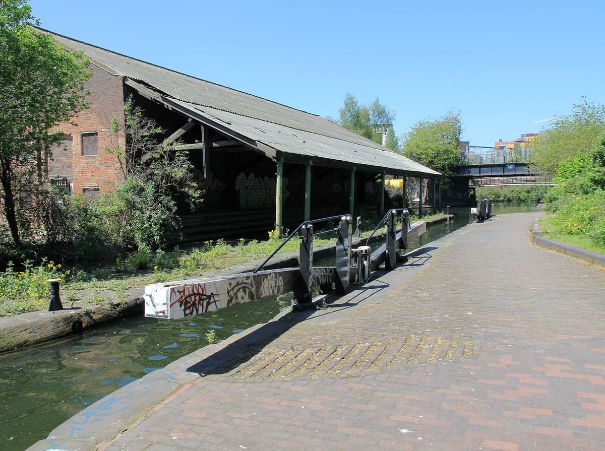 A canal stop lock with black-and-white balance beams beside a long brick warehouse whose roof overhangs the water on iron columns.