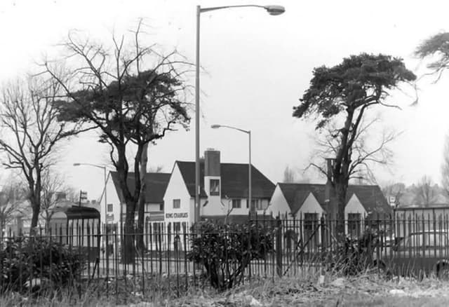 The white-painted King Charles pub with steep gables, seen through iron railings and trees, with street lamps in front