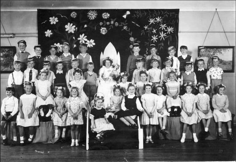An infant-school May Day group in a hall: a May Queen in a white dress and veil sits on a stepped throne in front of a large painted backdrop of daisies, with about forty children in party dresses, flower garlands and paper hats around her.