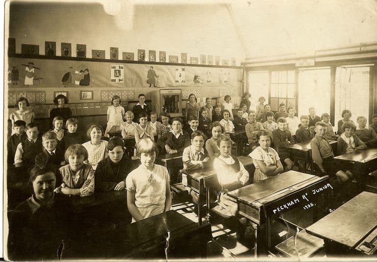 A sepia classroom photograph: about forty-five children at wooden double desks turn to face the camera, with children’s drawings and posters pinned along the wall and tall windows on the right. The front desk is chalked “Peckham Rd Junior 1935”.