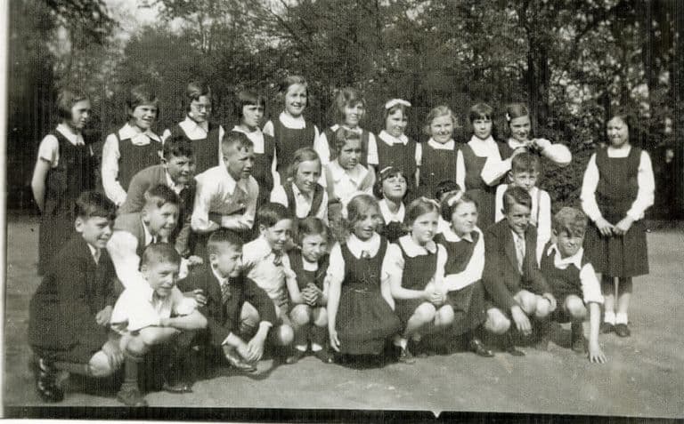 A class of about thirty boys and girls posed outdoors in front of trees, the girls standing behind in white blouses and dark pinafores and the boys crouching at the front.