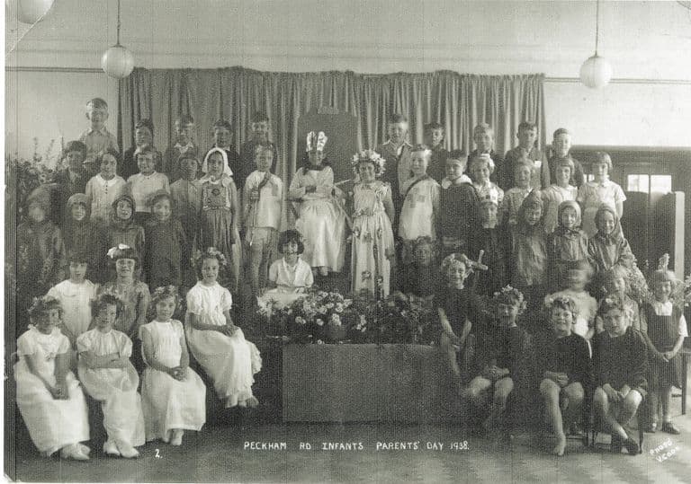 About fifty infants in costume on a stage in front of a curtain: a child in a crown sits on a throne among girls in white dresses with flower garlands, boys in tabards, and a bank of flowers at the front. The print is titled “Peckham Rd Infants Parents’ Day 1938”.
