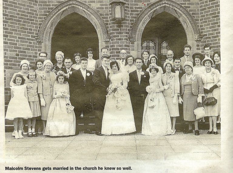 A newspaper photograph of a wedding party of about forty people in front of the church’s two pointed-arch brick doorways: the bride and groom in the centre with bridesmaids and flower girls in long dresses, guests in hats and suits. The printed caption reads “Malcolm Stevens gets married in the church he knew so well.”