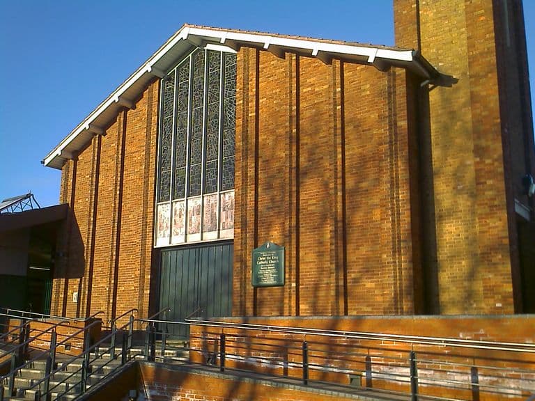 The brick front of Christ the King church in low winter sun: a tall gable with a full-height stained-glass window over green doors, a noticeboard, a square tower on the right and steps with handrails leading up.