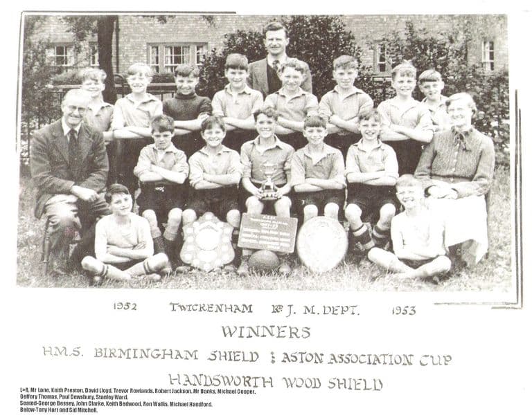A school football team of about fifteen boys with two masters and a woman teacher, posed on grass in front of a brick building with a cup, two shields and a ball; the mount reads “1952 Twickenham Rd J. M. Dept. 1953 – Winners H.M.S. Birmingham Shield, Aston Association Cup, Handsworth Wood Shield”, with the names typed below.