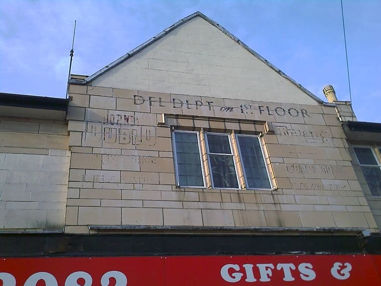 The pale stone gable of a shop building, with the ghost of old painted lettering around a leaded window: “DFL DEPT on 1st FLOOR”, “Hornby Dublo”, “Trains”, “Meccano” and “Corgi Toys” can still be made out. A red shop sign runs along the bottom.