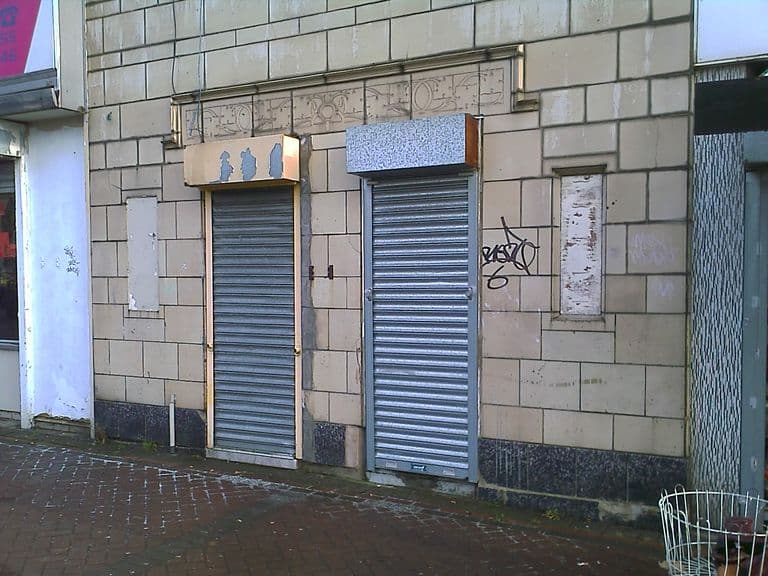 Two narrow doorways closed with metal roller shutters in a pale faience-tiled shop front, beneath a moulded frieze of stylised flowers; graffiti on the wall and a block-paved pavement in front.