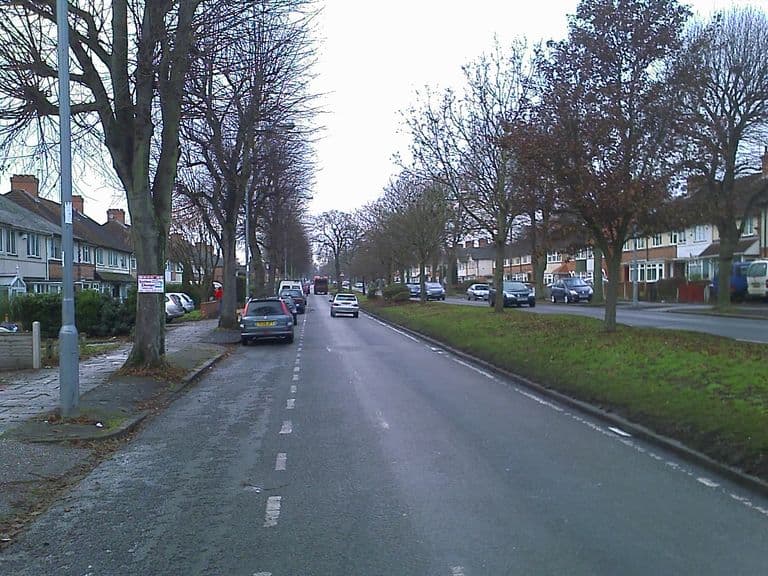 A long straight dual carriageway lined with bare trees and a wide grass central reservation, with semi-detached houses on both sides and parked cars along the kerb.