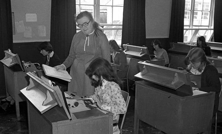 A black-and-white classroom scene: a teacher in glasses and a spotted dress stands holding a sheet of paper beside a pupil at a typewriter, while other girls work at typewriters on wooden desks with angled copy stands; tall windows behind.