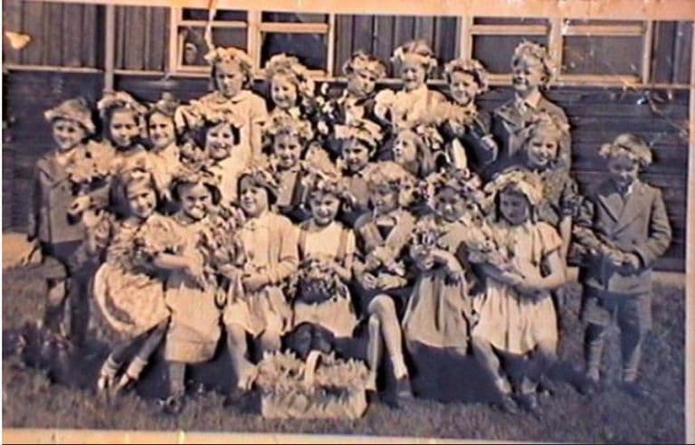 About thirty young children, most of the girls wearing flower garlands on their heads and holding flowers, crowded together on grass in front of a wooden school building; a basket of flowers sits at the front.
