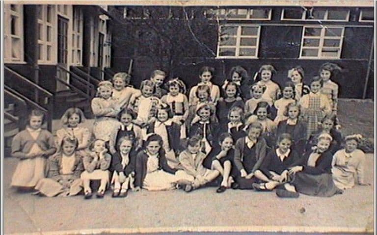 A class of girls, some sitting cross-legged on the playground and others standing behind, in front of the school’s wooden classroom blocks; a flight of wooden steps with handrails leads up to a classroom door on the left.