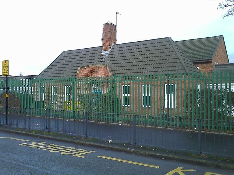 A single-storey red-brick school building with a tiled hipped roof and a tall chimney behind green palisade fencing, with “SCHOOL KEEP CLEAR” markings on the road in front.