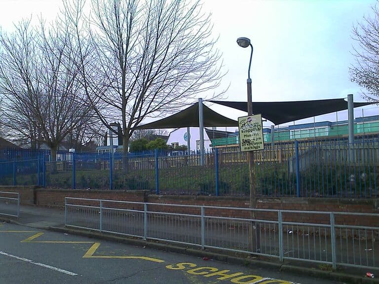 Modern school buildings with shade sails over the playground, seen behind blue railings and a low brick wall from Cranbourne Road; bare trees, a lamp post with a no-stopping sign and “SCHOOL” road markings in front.