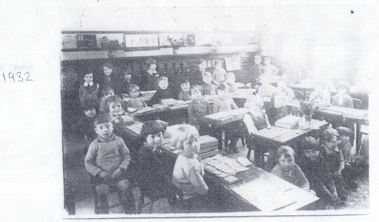 A faded photocopy of a classroom photograph: rows of small children at wooden desks with slates and papers, pictures along the wall and a vase of flowers; “1932” is handwritten beside it.