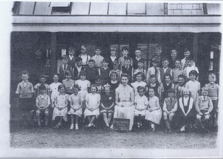 A class of about forty-five children in rows on grass in front of a verandah, with a woman teacher seated in the centre; a slate at her feet reads “Kingsthorne School”.