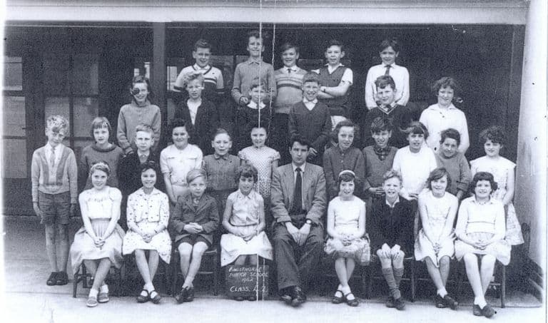 A class of about thirty-five children in four rows with a young male teacher seated in the centre, under a verandah; a slate at the front reads “Kingsthorne Junior School 1962 Class 4.2”.
