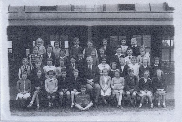A class of about forty children in four rows with a male teacher seated in the centre, in front of a verandah and windows; a boy at the front holds a slate reading “Kingsthorne School”.