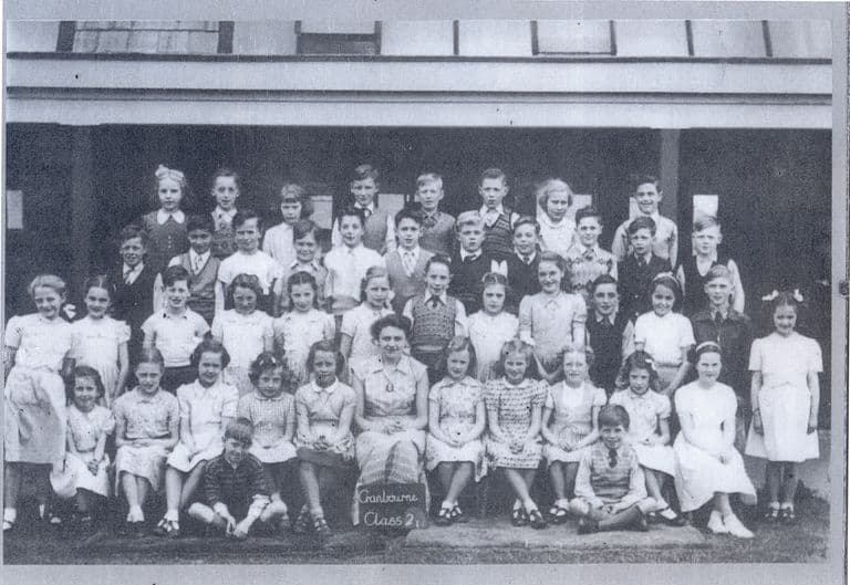 A class of about forty-five children in rows with a woman teacher seated in the middle, the girls in summer dresses; a slate at the front reads “Cranbourne Class 2”.