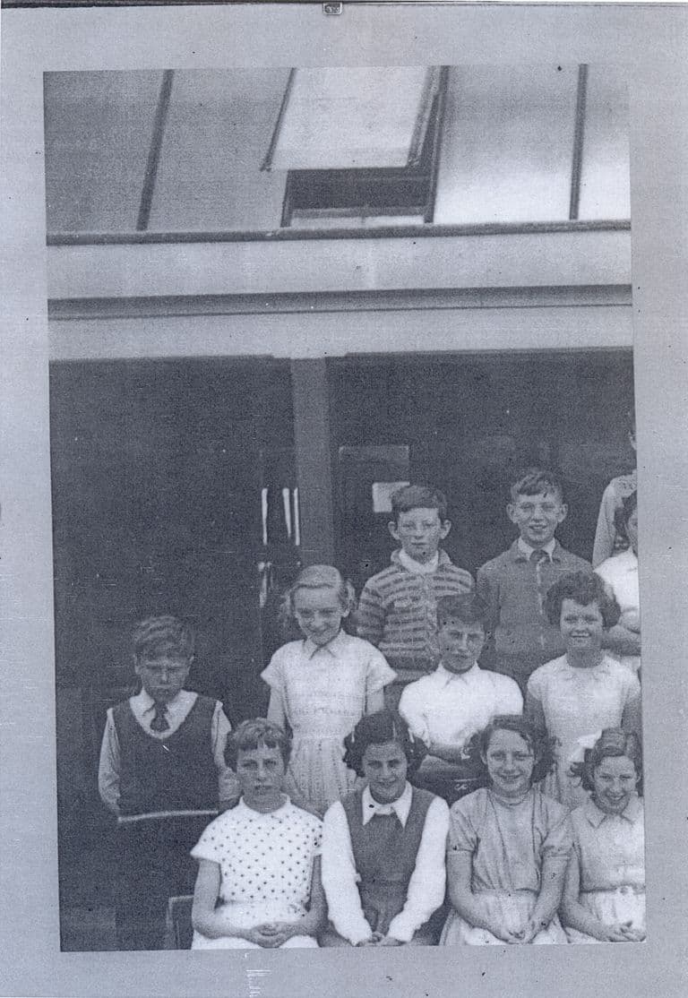 Part of a class photograph mounted on a grey album page: about a dozen children in three rows in front of a verandah and roof light, the rest of the class cut off at the left.