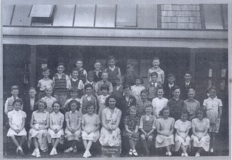 A class of about forty children in rows with a woman teacher seated in the centre, in front of a verandah with roof lights; a slate at the front reads “Kingsthorne School”.