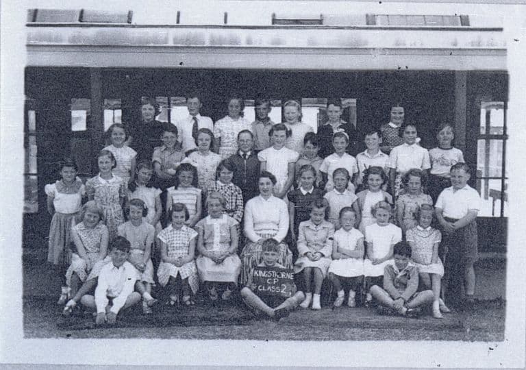 A class of about forty children in rows with a woman teacher in the centre, girls in patterned summer dresses; a boy at the front holds a slate reading “Kingsthorne C.P. Class 2”.