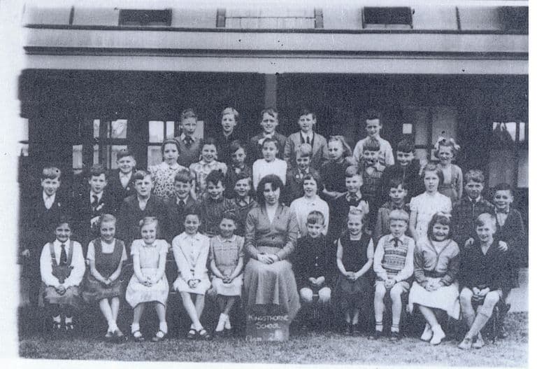 A class of about forty children in rows in front of a verandah, with a woman teacher seated in the centre; a slate at her feet reads “Kingsthorne School”.